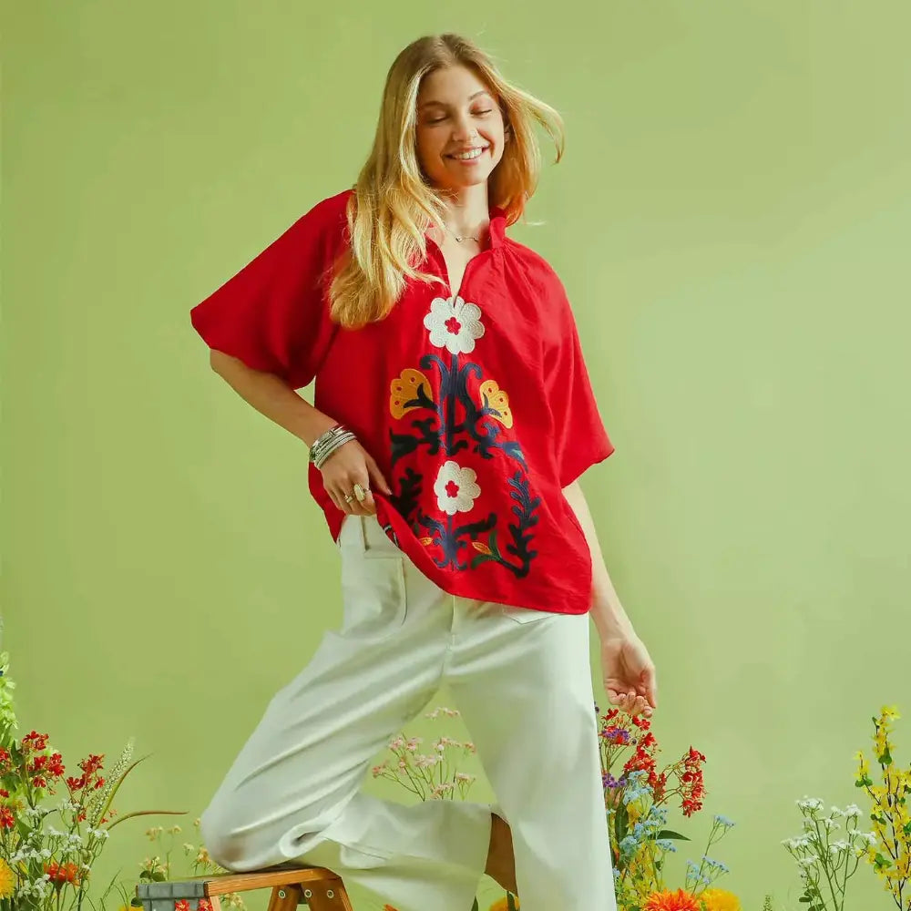 A smiling woman wearing a vibrant red oversized button-up shirt with large white floral embroidery, paired with white trousers, posing in a field of wildflowers.