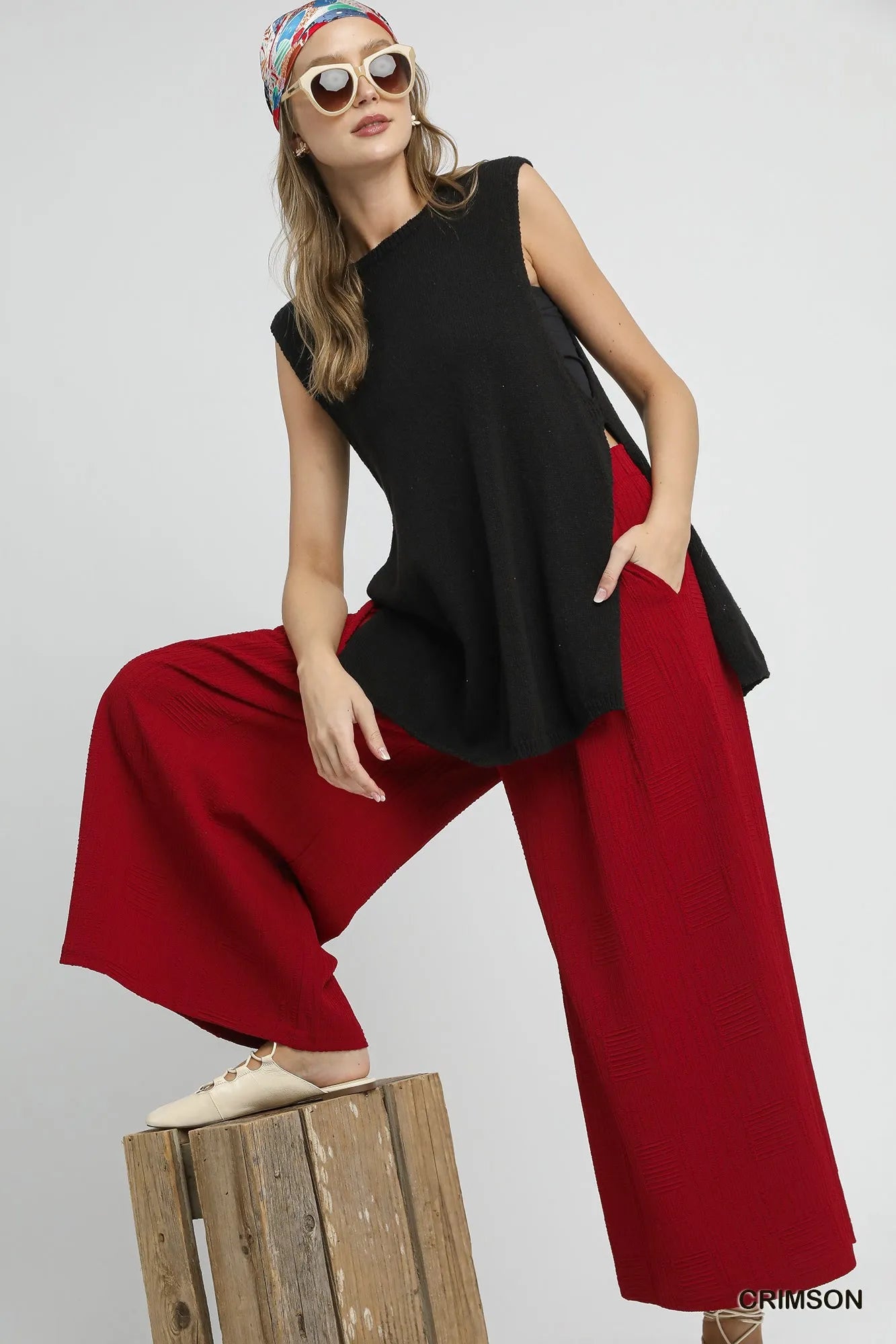 Woman wearing a black sleeveless top and red pants sitting on a wooden block against a gray background.