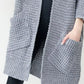 Woman wearing a cozy, longline gray chunky knit open-front cardigan with a textured waffle weave and large front pockets.