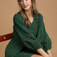 Woman wearing a green dress with brown trim sitting on a red chair against a beige background