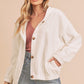 Woman wearing a white jacket over a pink top and denim shorts on a beige background