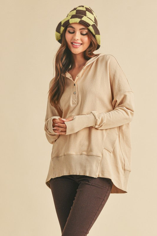 Woman wearing a beige long-sleeve hoodie, brown pants, and a patterned hat on a beige background