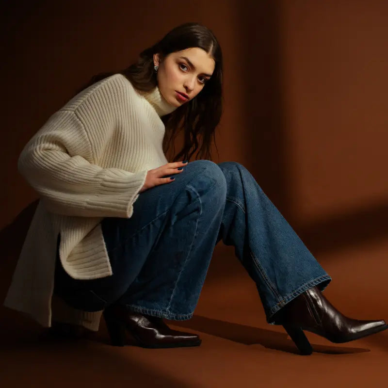 Woman wearing a white sweater, blue jeans, and black boots sitting on a brown background