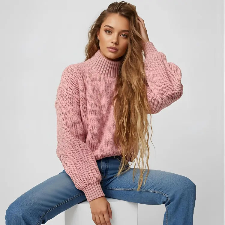 Woman wearing a pink knitted sweater and blue jeans sitting on a white stool against a light gray background