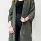 Woman wearing a longline olive green chunky knit open-front cardigan with a textured waffle weave and deep front pockets, styled over a black outfit.