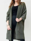 Woman wearing a longline olive green chunky knit open-front cardigan with a textured waffle weave and deep front pockets, styled over a black outfit.