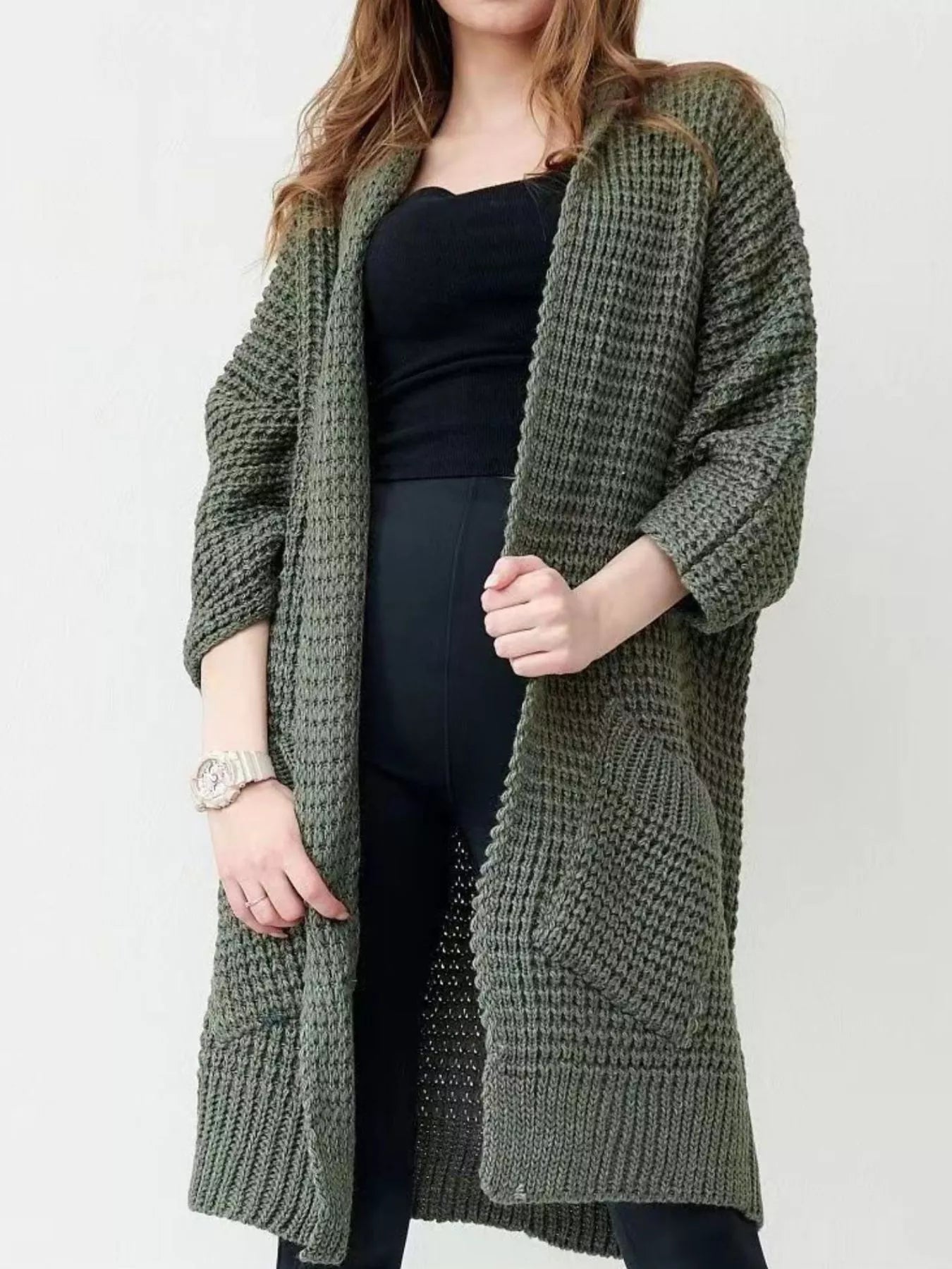 Woman wearing a longline olive green chunky knit open-front cardigan with a textured waffle weave and deep front pockets, styled over a black outfit.