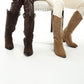 Two women wearing a pair of knee-high boots, one brown with studs and one beige, on a white background.