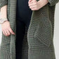 Woman wearing a longline olive green chunky knit open-front cardigan with a textured waffle weave and deep front pockets, styled over a black outfit.