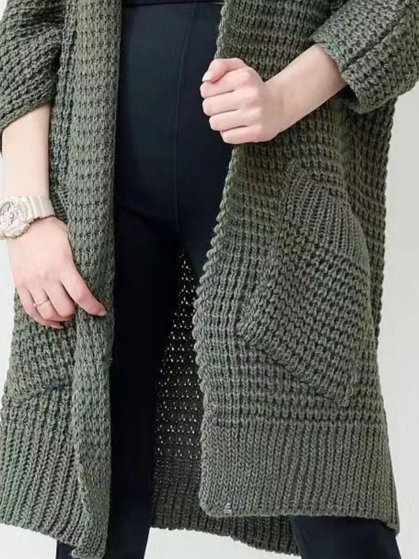 Woman wearing a longline olive green chunky knit open-front cardigan with a textured waffle weave and deep front pockets, styled over a black outfit.