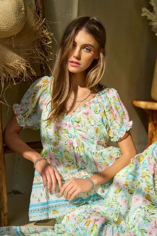 A woman wears a light blue and pink floral print dress with ruffled short sleeves, lace trim at the waistband, and delicate embroidery details.