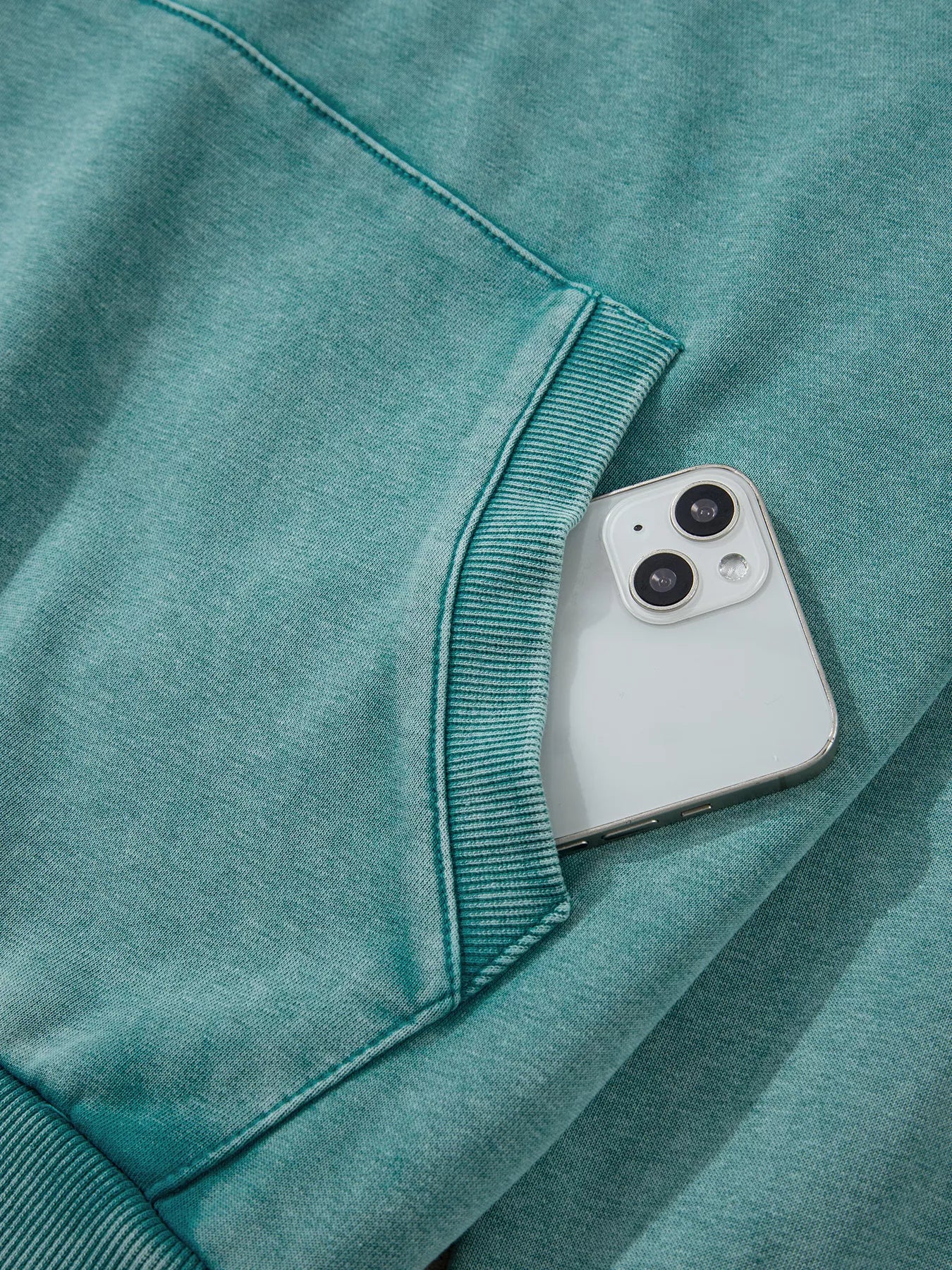 White iPhone 13 peeking from pocket of teal mineral wash hoodie