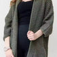 Woman wearing a longline olive green chunky knit open-front cardigan with a textured waffle weave and deep front pockets, styled over a black outfit.