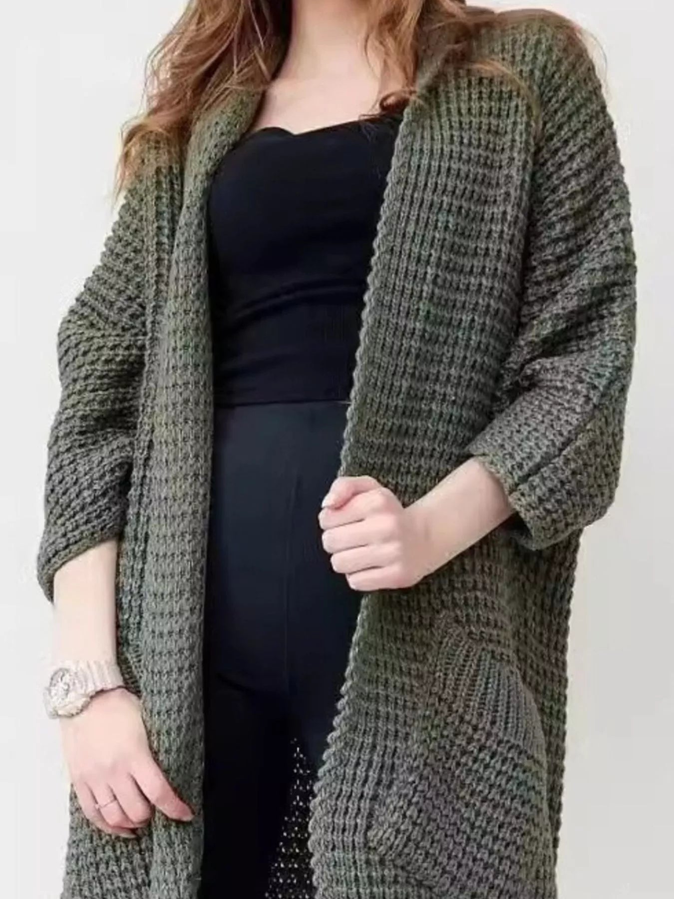 Woman wearing a longline olive green chunky knit open-front cardigan with a textured waffle weave and deep front pockets, styled over a black outfit.