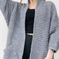 Woman wearing a cozy, longline gray chunky knit open-front cardigan with a textured waffle weave and large front pockets.