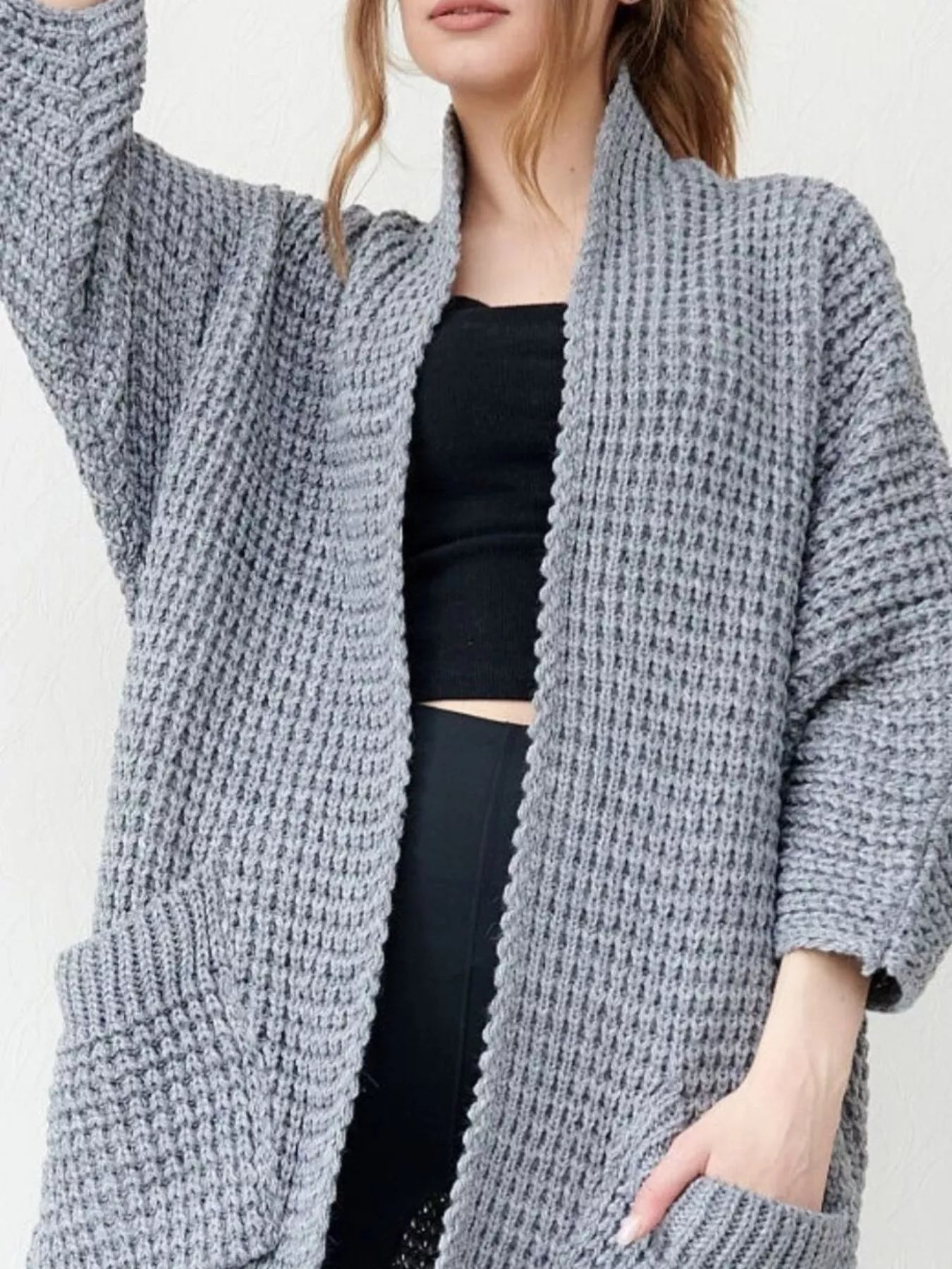 Woman wearing a cozy, longline gray chunky knit open-front cardigan with a textured waffle weave and large front pockets.