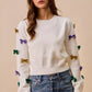 White knit sweater with colorful sequined bows in gold, purple, and green
