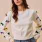 White knit sweater with colorful sequined bow embellishments on sleeves