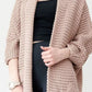 Woman wearing a blush-pink Chunky Knit Open Front Cardigan with a textured waffle weave and deep front pockets, styled over a black top.