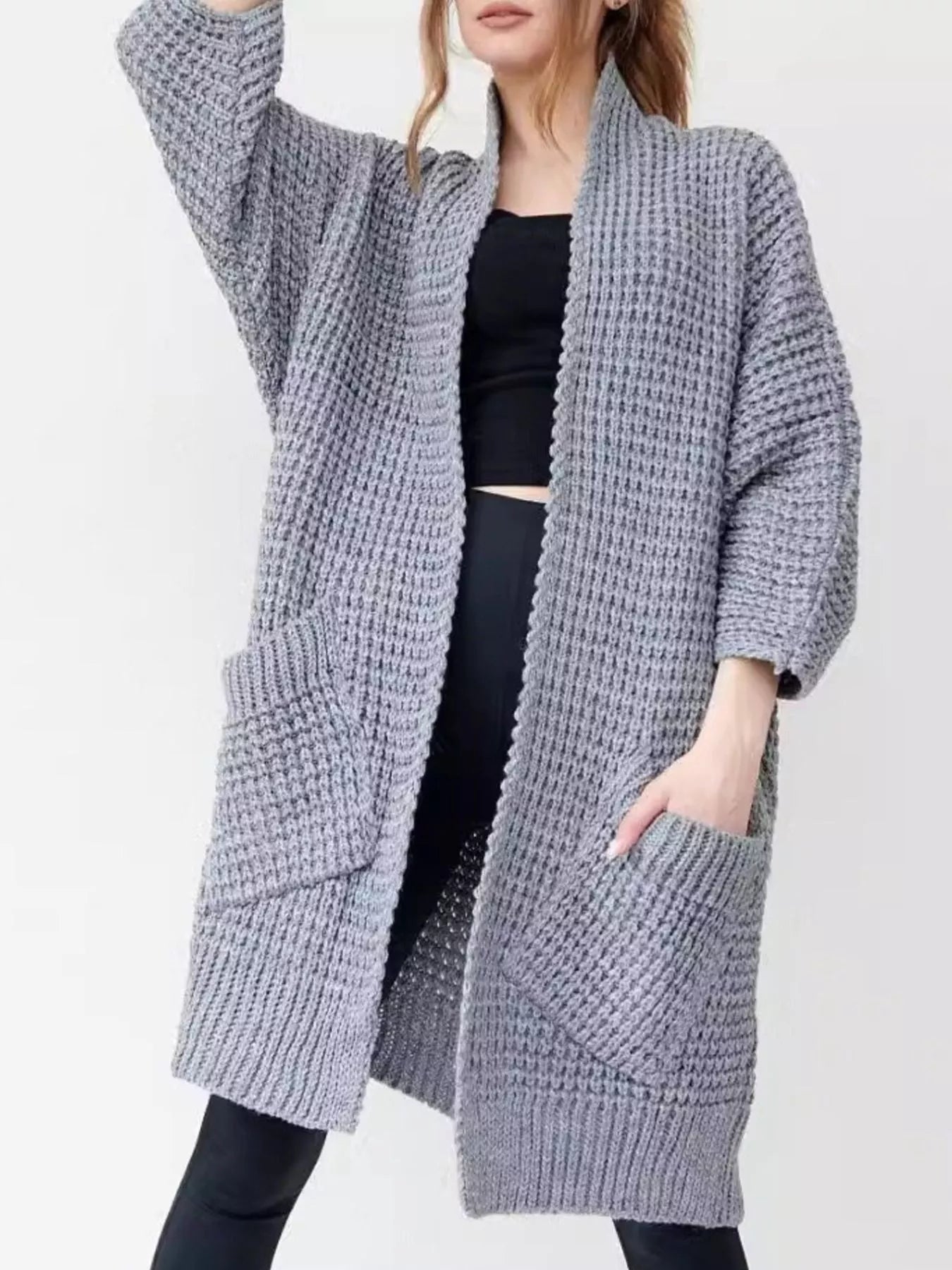 Woman wearing a cozy, longline gray chunky knit open-front cardigan with a textured waffle weave and large front pockets.